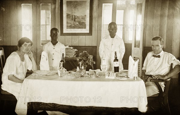 The Tamlins at table. Mr Alfred Tamlin and Mrs Dorothy Tamlin pose for the camera, sitting at a table with two African servants behind them. Gold Coast (Ghana), circa 1918. Ghana, Western Africa, Africa.