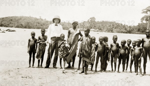 Children on the beach. A European stands on a beach surrounded by African children. Western Africa, circa 1920., Western Africa, Africa.