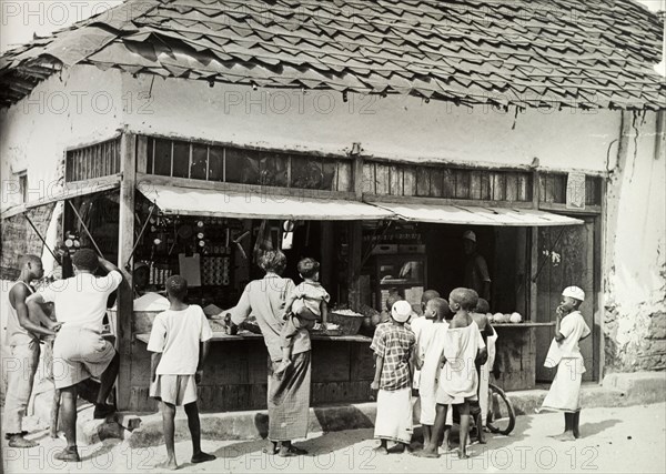 Tanganyika street shop. People wait to be served from a busy market stall housed in a ramshackle building. Tanganyika Territory (Tanzania), circa 1960. Tanzania, Eastern Africa, Africa.