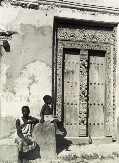 Carved wooden door, Zanzibar. A young man and a child sit in the sunshine beside an ornately carved wooden door. Zanzibar, Tanganyika (Tanzania), circa 1960. Zanzibar Town, Zanzibar Urban/West, Tanzania, Eastern Africa, Africa.