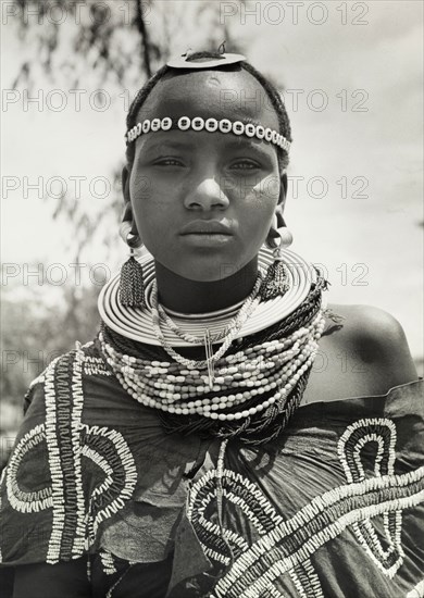 Young Maasai woman. Close-up portrait of a young Maasai woman wearing traditional jewellery including earrings, beaded necklaces and a beaded headband. Her wraparound robe is also adorned with beads sewn on in decorative patterns. Tanganyika Territory (Tanzania), circa 1960. Tanzania, Eastern Africa, Africa.
