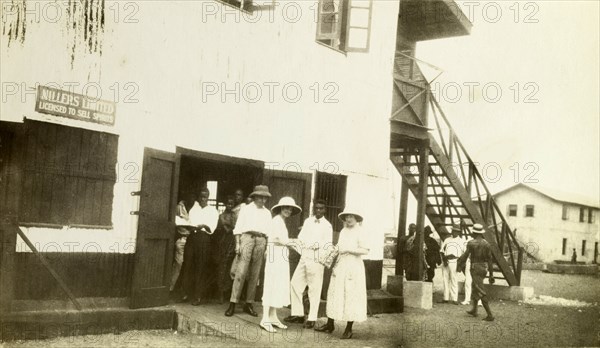 Millers Limited trading centre. Europeans and Africans outside the trading centre of Millers Limited. The sign near the door reads 'MILLERS LIMITED, LICENSED TO SELL SPIRITS'. Probably Accra, Gold Coast (Ghana), circa 1918. Ghana, Western Africa, Africa.