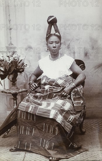Woman with an elaborate hairstyle, Gold Coast. Studio portrait of a smartly dressed African woman who wears one or two strip-woven cloth wraps (possibly kente cloth) together with a tailored blouse. Her hair has been fashioned in two steep cylindrical pillars which join together above her head in a sphere. She wears gold bracelets and rings and other jewellery, and holds a furled umbrella. Probably Accra, Gold Coast (Ghana), circa 1918. Ghana, Western Africa, Africa.