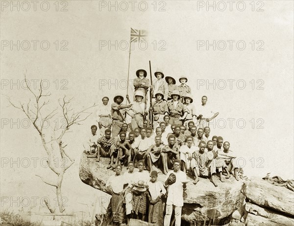 The flag on the rock. A union jack flag flies above a group of African and European Miller Brothers' employees who pose for the camera atop a rocky outcrop. Amongst the European men are Mr Alfred Tamlin and his colleague Mr Jackson. Western Africa, circa 1920., Western Africa, Africa.
