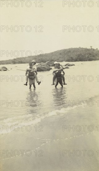 Out of water. Two European men wearing solatopi hats are carried above the water on the backs of two African men smoking cigarettes. Possibly Ghana, Western Africa, circa 1920., Western Africa, Africa.
