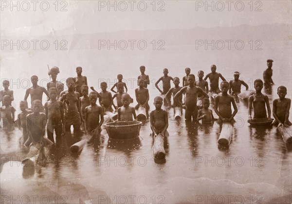 Children at lake shore, Gold Coast. A group of naked African people, mainly children, pose for the camera at a lake near Kumasi. Some of them sit astride floating logs, possibly a fishing aid or perhaps a recreational toy. Gold Coast (Ghana), circa 1920., Ashanti, Ghana, Western Africa, Africa.