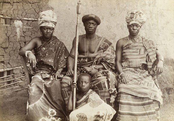 The Mensa of Kumasi and his family. Outdoors portrait of the Mensa of Kumasi with his two wives and a son. He is shown here wearing a kente robe, as does his wife on the right. The wife on the left wears one or two wraps of tie-dye fabric, while the boy wears a robe of printed cotton. The child holds a long metal staff, possibly his father's staff of office. Kumasi, Gold Coast (Ghana), circa 1920. Kumasi, Ashanti, Ghana, Western Africa, Africa.