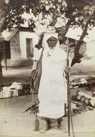 Curio seller at Accra. Outdoors portrait of an aged and apparently well-known itinerant vendor, said to be named 'Arrabi', balancing bundles of cloth on his head. Gold Coast (Ghana), 1917. Ghana, Western Africa, Africa.
