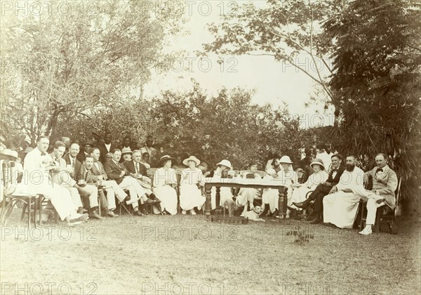Miller Brothers agency staff outing. European employees of Miller Brothers agency are seated outdoors around a table with wives and friends, including a priest. Some African employees or servants are standing in the background. Probably Accra, Gold Coast (Ghana), circa 1920. Ghana, Western Africa, Africa.
