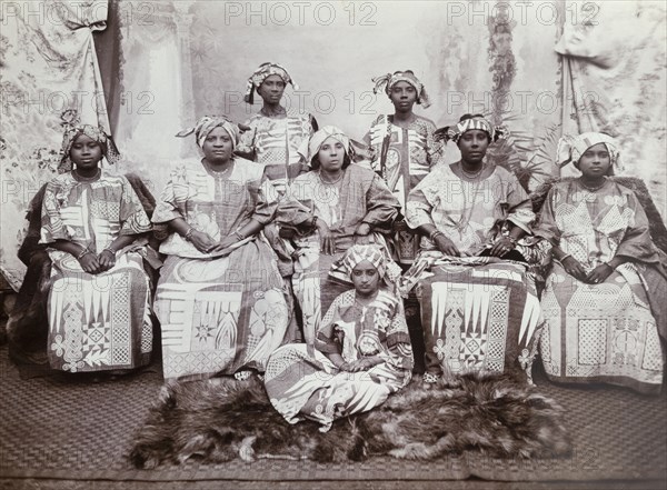 Eight women in indigo dresses. The women wear tailored gowns of dark cloth (probably indigo blue cotton) embroidered with geometrical patterns, suggesting that they are Hausa women from northern Nigeria. They also wear head cloths. Western Africa, circa 1918., Western Africa, Africa.