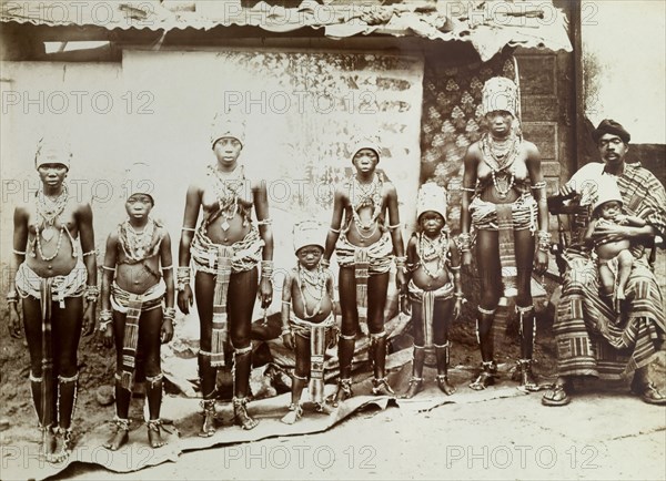 Women in beaded costume. Outdoors portrait of girls and young women wearing elaborate beaded and strip-cloth aprons, tall headdresses and heavy jewellery, with a seated man wearing kente cloth and a baby. Gold Coast (Ghana), circa 1920. Ghana, Western Africa, Africa.