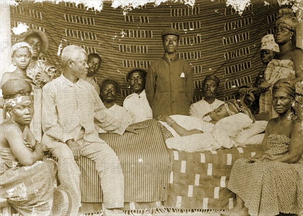 Grieving family, Western Africa. A family surround the body of a deceased female relative, lying covered with sheets on a bed. Western Africa, circa 1920., Western Africa, Africa.