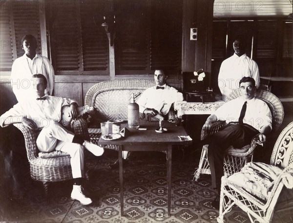 European staff relaxing in Swanzy's bungalow. Alfred Tamlin, A. M. Napolitano, and P. Kane relax in the managers' bungalow of the Motor Transport Depot of F. & A. Swanzy Limited. Behind them stand two African servants, both men. Accra, Gold Coast (Ghana), April 1918. Accra, East (Ghana), Ghana, Western Africa, Africa.