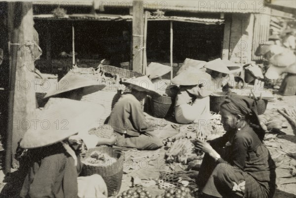 Chinese market in Burma (Myanmar). Shan traders, dressed in traditional conical hats, sit on the ground amongst their wares at a Chinese bazaar in Mogok. Mogok, Burma (Myanmar), circa 1925. Mogok, Sagaing, Burma (Myanmar), South East Asia, Asia.