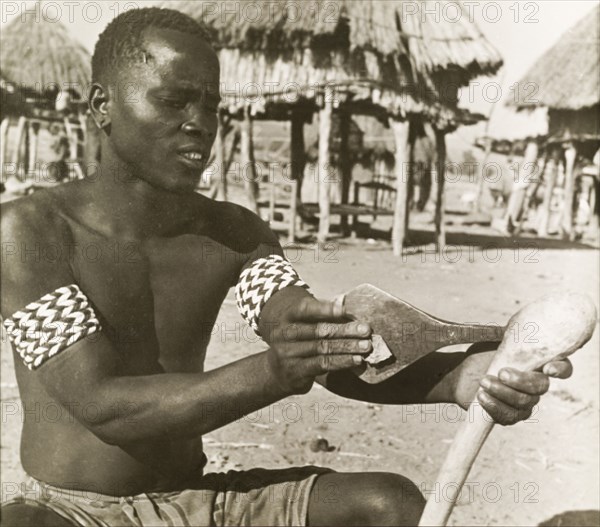 A Tonga man sharpens an axe. A semi-naked Tonga (Batonga) man wearing patterned armbands sharpens the blade of a wooden-handled axe. Probably Southern Rhodesia (Zimbabwe), circa 1955. Zimbabwe, Southern Africa, Africa.