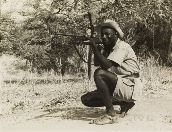 Taking aim during a film shoot. An African man wearing sandals and a cowboy hat, crouches down on one knee to take aim with his rifle. This photograph was taken during a film shoot for the Central African Film Unit (CAFU) and may feature an actor or crew member involved with the film. Probably Southern Rhodesia (Zimbabwe), circa 1955. Zimbabwe, Southern Africa, Africa.
