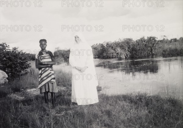 Mother Superior and student. The Mother Superior of the Chilabula Mission, dressed in white from head to toe, stands beside one of her female African students on the banks of a river. Near Kasama, Northern Rhodesia (Zambia), circa 1952., North (Zambia), Zambia, Southern Africa, Africa.