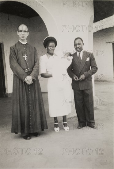After the christening. A proud African couple pose beside a European priest following the christening of their baby. Northern Rhodesia (Zambia), circa 1952. Zambia, Southern Africa, Africa.