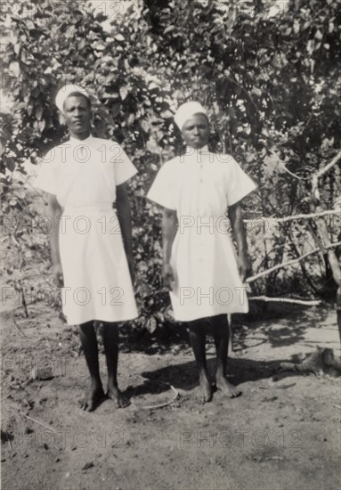 Cigarette and Timothy'. Two domestic servants, identified by an original caption as 'Cigarette and Timothy', pose for the camera. Their pristine uniform consists of a knee-length white dress, belted at the waist and worn with a starched white hat. Northern Rhodesia (Zambia), circa 1952. Zambia, Southern Africa, Africa.