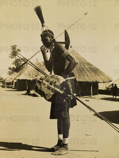 Tonga man in mourning dress. Full-length profile portrait of a Tonga man wearing traditonal mourning dress. He wears an upright tassled headdress and carries several spears and a shield. Gwembe, Northern Rhodesia (Zambia), circa 1950. Gwembe, South (Zambia), Zambia, Southern Africa, Africa.