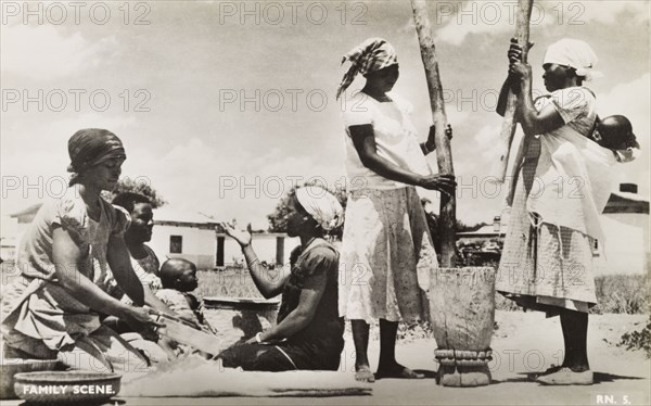 A domestic scene. Domestic scene showing a group of women working together outdoors in an African village. One woman separates grain with a sieve, whilst two others pound it using a large pestle and mortar. Southern Africa, circa 1950., Southern Africa, Africa.