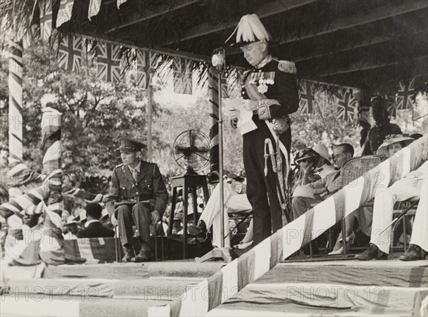 Arden-Clarke delivers a speech. Sir Charles Noble Arden-Clarke (1898-1962), last Governor of the Gold Coast and first Governor General of independent Ghana, steps up to the microphone to deliver a speech on an outdoor stage decorated with union jack flags. Accra, Gold Coast (Ghana), circa 1950. Accra, East (Ghana), Ghana, Western Africa, Africa.