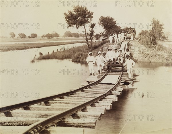 The pontoon line. A section of railway line stretches across a flooded river, supported only by a makeshift pontoon that sits in the swollen waters. Three men sit on a hand-operated rail cart, preparing to cross, whilst a full-sized steam engine waits in the distance. West Bengal, India, circa 1920., West Bengal, India, Southern Asia, Asia.