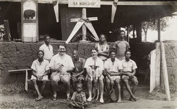 Workshop employees. Two British colonial officers identified as Charles Simpson (left) and Cyril Iles (right), sit on a bench outside a wood workshop with several African men. Woodworking tools including saws and planes are proudly arranged beneath a sign that reads: 'N A Workshop'. Okitipupa, Nigeria, circa 1945. Okitipupa, Ondo, Nigeria, Western Africa, Africa.