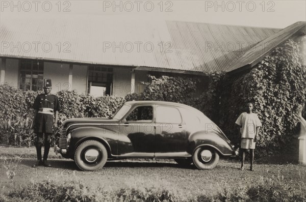 Servants to a colonial officer. A uniformed orderly serving as an attendant to a British colonial officer and a domestic servant identified as 'Gabriel', stand beside a small car parked in a driveway. Southern Nigeria, circa 1950. Nigeria, Western Africa, Africa.