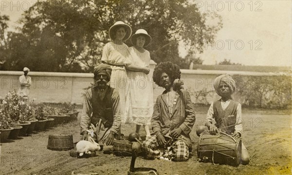 A sacrificial rabbit. Two European women stand fixated, watching as a group of snake charmers present their cobra with a sacrificial rabbit. Bangalore, India, 1922. Bangalore, Karnataka, India, Southern Asia, Asia.