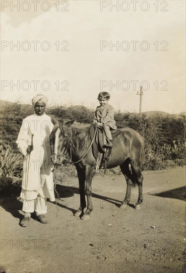 An Indian 'syce'. An Indian 'syce' (hostler) guides a pony ridden by a young British child along a country lane. Bombay (Mumbai), India, circa 1920. Mumbai, Maharashtra, India, Southern Asia, Asia.