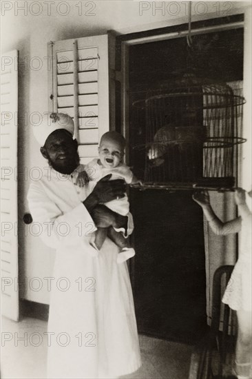 Beside the parrot's cage'. An Indian servant poses for the camera holding a British baby who seems greatly entertained by the household's caged parrot. Calcutta (Kolkata), India, 1932. Kolkata, West Bengal, India, Southern Asia, Asia.