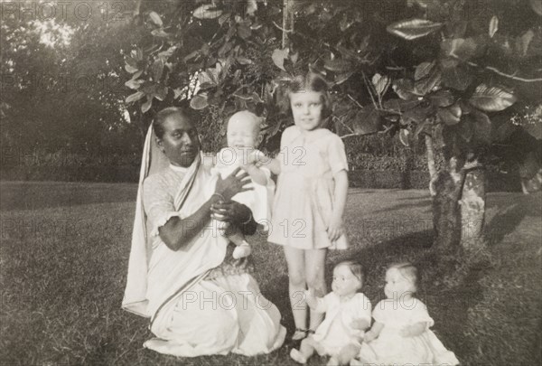Ayah with children. An Indian ayah (nursemaid) poses beside her British charges, a young girl and a baby, in the garden of a colonial residence. Two toy dolls are propped up for the camera in the foreground. Calcutta (Kolkata), India, October 1932. Kolkata, West Bengal, India, Southern Asia, Asia.