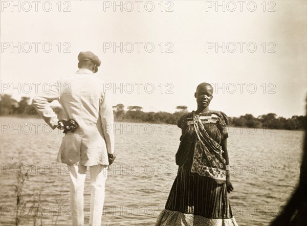 An African man and woman at the Zambezi River. Portrait of an African man and woman on the banks of the Zambezi River. The woman faces the camera, traditionally dressed in patterned clothing, whilst the man shows his back, dressed in a smart, Western-style white suit. Southern Rhodesia (Zimbabwe), circa 1930. Victoria Falls, Matabeleland North, Zimbabwe, Southern Africa, Africa.
