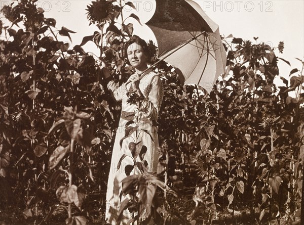 Amongst the sunflowers at Balloch Farm. A European woman, wearing a striped dress, shades herself with a parasol as she stands in a field of sunflowers that reach higher than her head at Balloch Farm. Near Gwelo, Southern Rhodesia (near Gweru, Zimbabwe), circa 1930. Gweru, Midlands, Zimbabwe, Southern Africa, Africa.