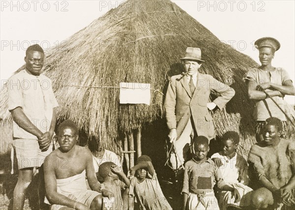 Portrait of workers and children at Balloch Farm. Portrait of a group of male African farm workers, assembled outdoors beside a round, thatched hut at Balloch Farm. They pose with four children and a European man, probably an overseer, who wears a suit and smokes a pipe. Near Gwelo, Southern Rhodesia (near Gweru, Zimbabwe), circa 1930. Gweru, Midlands, Zimbabwe, Southern Africa, Africa.