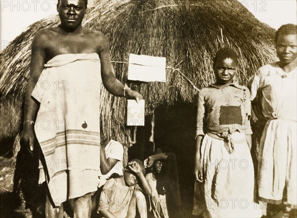 African worker with children at Balloch Farm. An African farm worker holds up a piece of paper or book for the camera, whilst posing with several children outside a round, thatched hut at Balloch Farm. Near Gwelo, Southern Rhodesia (near Gweru, Zimbabwe), circa 1930. Gweru, Midlands, Zimbabwe, Southern Africa, Africa.