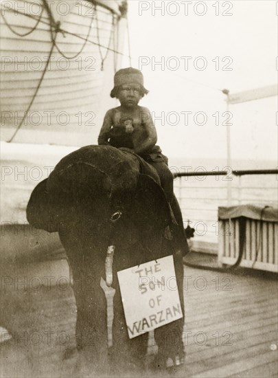 Son of Warzan'. A small boy dressed as Tarzan rides on the back of a stuffed toy elephant, clutching a toy monkey in his lap during a children's fancy dress party aboard RMS Arundel Castle, enroute from southern Africa to England. A sign strung around the elephant's neck reads: 'The Son of Warzan'. Location unknown, circa 1930.