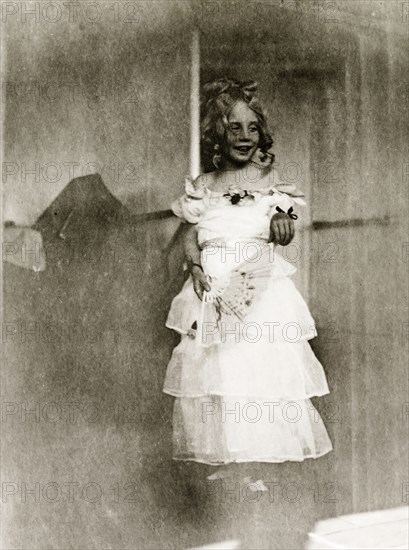 Girl in fancy dress aboard RMS Arundel Castle. Portrait of a young girl dressed in costume for a children's fancy dress party aboard RMS Arundel Castle, enroute from southern Africa to England. Her multi-tiered, strapless party dress is tied at the waist with a sash, her hair is worn up and she carries a decorative fan in one hand. Location unknown, circa 1930.