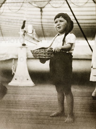 Boy in fancy dress aboard RMS Arundel Castle. Portrait of a young boy dressed in costume for a children's fancy dress party aboard RMS Arundel Castle, enroute from southern Africa to England. Barefooted, he wears a shirt and shorts, and supports a basket, which is strung around his neck. Location unknown, circa 1930.