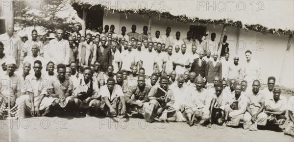 A Methodist mission in the Tana River area. A number of African men pose for a group portrait outside a low, thatched mission building in the Tana River area. English Methodist missionary, Reverend Ian Lewis, stands at the far right in the middle row. Coast Province, Kenya, August 1952., Coast, Kenya, Eastern Africa, Africa.