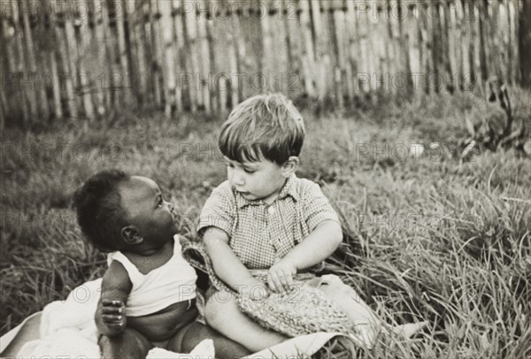 Anthony Lewis befriends an African baby. Anthony Lewis, a youngest son of English Methodist missionary, Reverend Ian Lewis, sits beside an African baby on a patch of grass outside the Lewis family home. Limuru, Kenya, circa 1956. Limuru, Central (Kenya), Kenya, Eastern Africa, Africa.