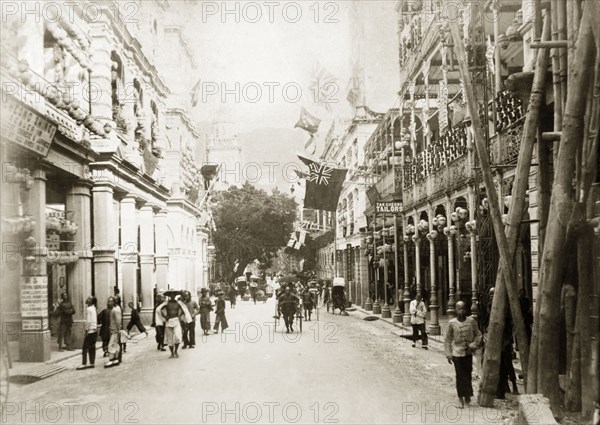 Queen's Road Central, Hong Kong. Rickshaws, pedestrians and street traders bustle about on Queen's Road Central. The Pedder Street clock tower, demolished in 1908, is visible in the distance: an original caption comments that it is covered 'with bamboo scaffolding for repairs'. Hong Kong, China, April 1903. Hong Kong, Hong Kong, China, People's Republic of, Eastern Asia, Asia.
