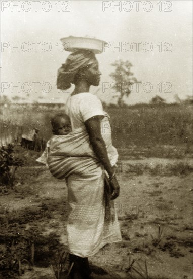 Nigerian woman and baby in profile. Profile portrait of a Nigerian women, identified by an original caption as 'Dick's wife'. She carries a small baby strapped to her back and balances a flat bowl on her head. Abeokuta, Ogun State, Nigeria, circa 1929. Abeokuta, Ogun, Nigeria, Western Africa, Africa.