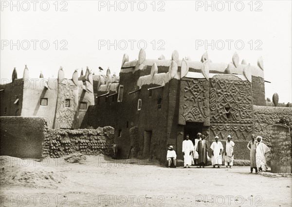 An ancient Hausa building in Kano. A number of Nigerian men stand outside a mud-walled building, constructed in traditional Hausa style and decorated with geometric carvings. This is probably the Emir's Palace, built by Sarkin Mohammadu Rumfa (1463-99) in the fifteenth century. Kano, Kano State, Nigeria, circa 1930. Kano, Kano, Nigeria, Western Africa, Africa.
