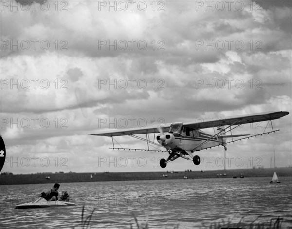 Airspray demonstration. A small plane flies low over the water at the Aquasports Regatta. Kenya, 28 October 1956. Kenya, Eastern Africa, Africa.