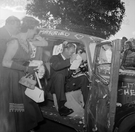 Just married. Guests crowd around the newlywed Carey and Courtney couple as they dive into their wedding car covered with graffiti from well-wishers. Kenya, 15 December 1956. Kenya, Eastern Africa, Africa.