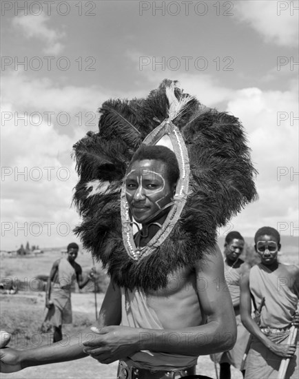 Wakamba dancer in headdress. Portrait of a male Wakamba dancer, his face painted and wearing a large, mane-like headdress made from black feathers for Princess Margaret's visit. Machakos, Kenya, 22 October 1956. Machakos, East (Kenya), Kenya, Eastern Africa, Africa.