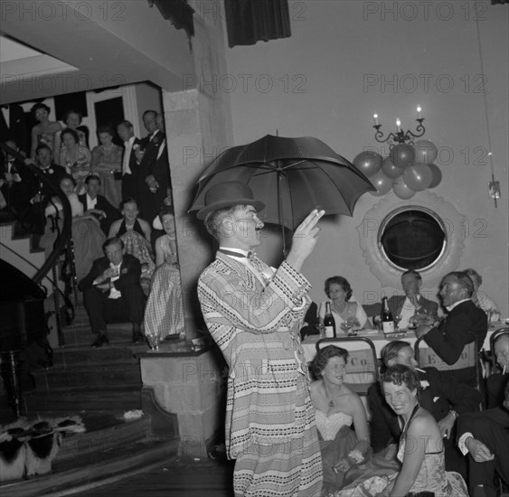 Cabaret act at the Limuru hunt ball. A cabaret performer wearing a flamboyant suit and holding an umbrella, entertains a crowd of party guests at the Limuru hunt ball. Limuru, Kenya, 27 October 1956. Limuru, Central (Kenya), Kenya, Eastern Africa, Africa.