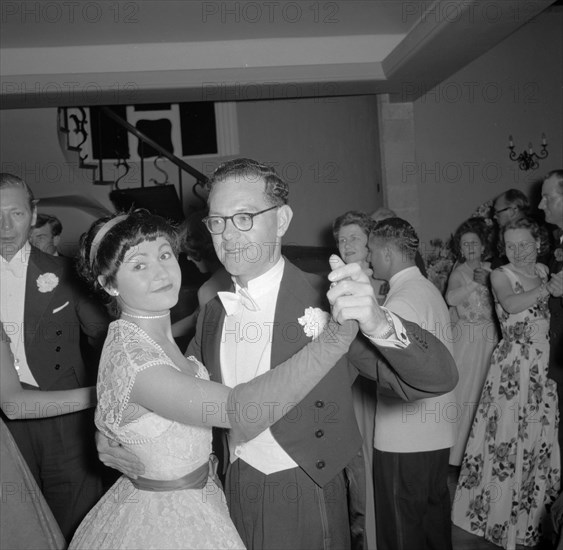 Guests at the Limuru hunt ball. A formally dressed European couple dance together at the Limuru hunt ball. The woman wears a white lace dress with long gloves, the man, a dinner jacket and bow tie. Limuru, Kenya, 27 October 1956. Limuru, Central (Kenya), Kenya, Eastern Africa, Africa.